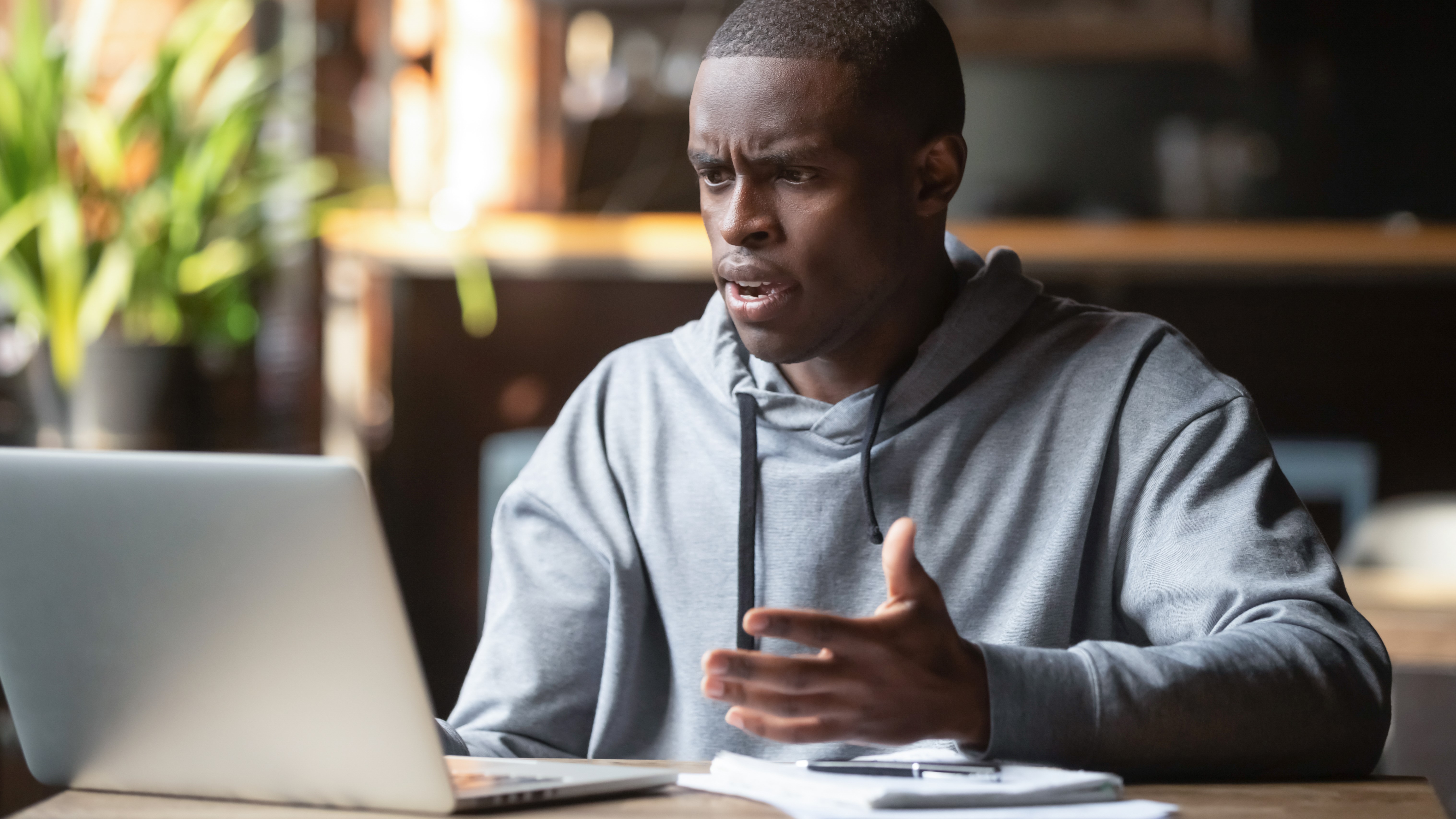 A man on a desk using a laptop and holding his hands up while he has a confused look on his face