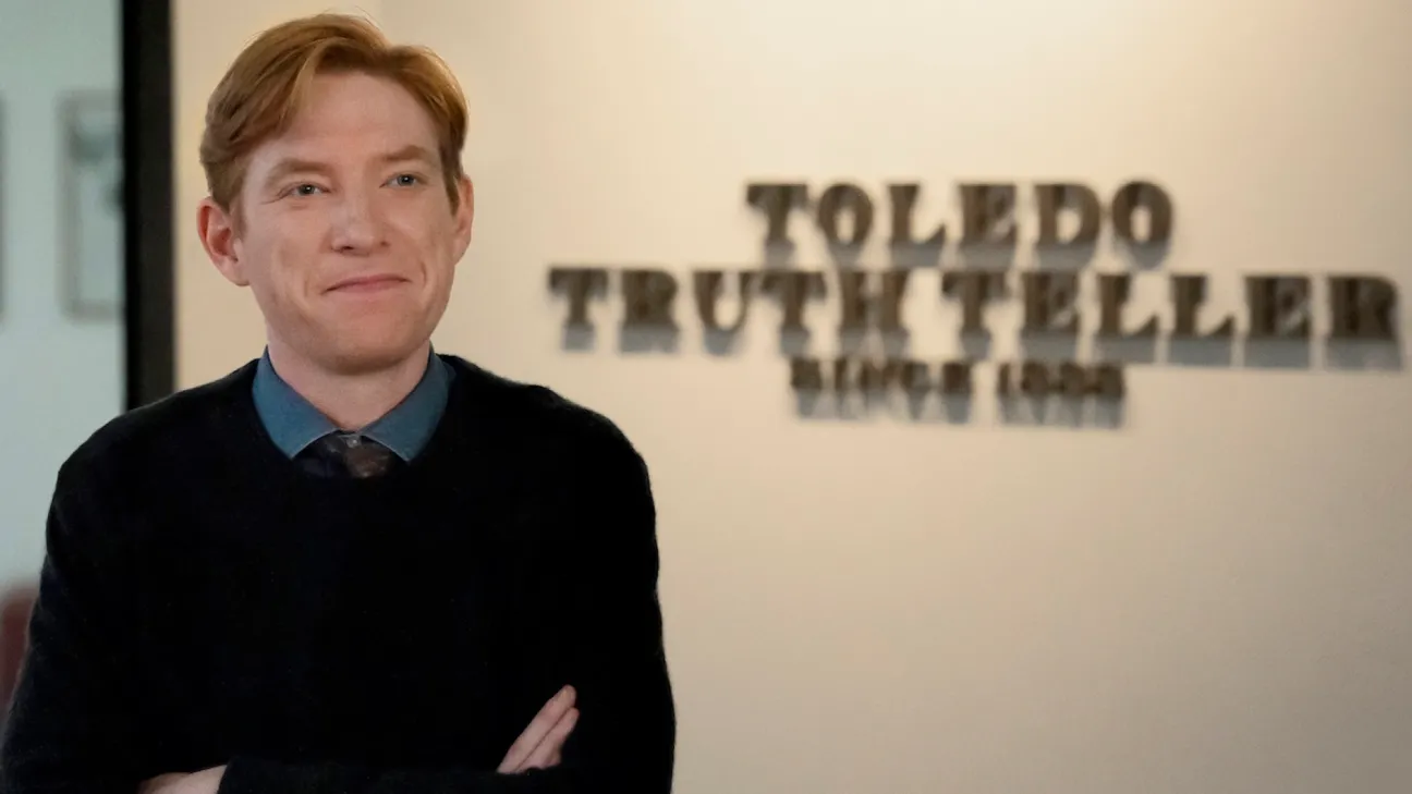 Domnhall Gleeson stands in front of a sign for the newspaper Toledo Truth Teller