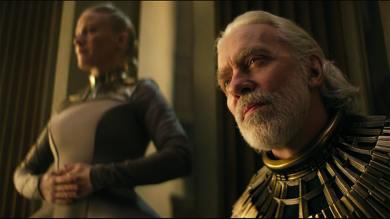 Brother Dusk is sitting down with an out-of-focus Demerzel nearby in Foundation Season 3 Section 8