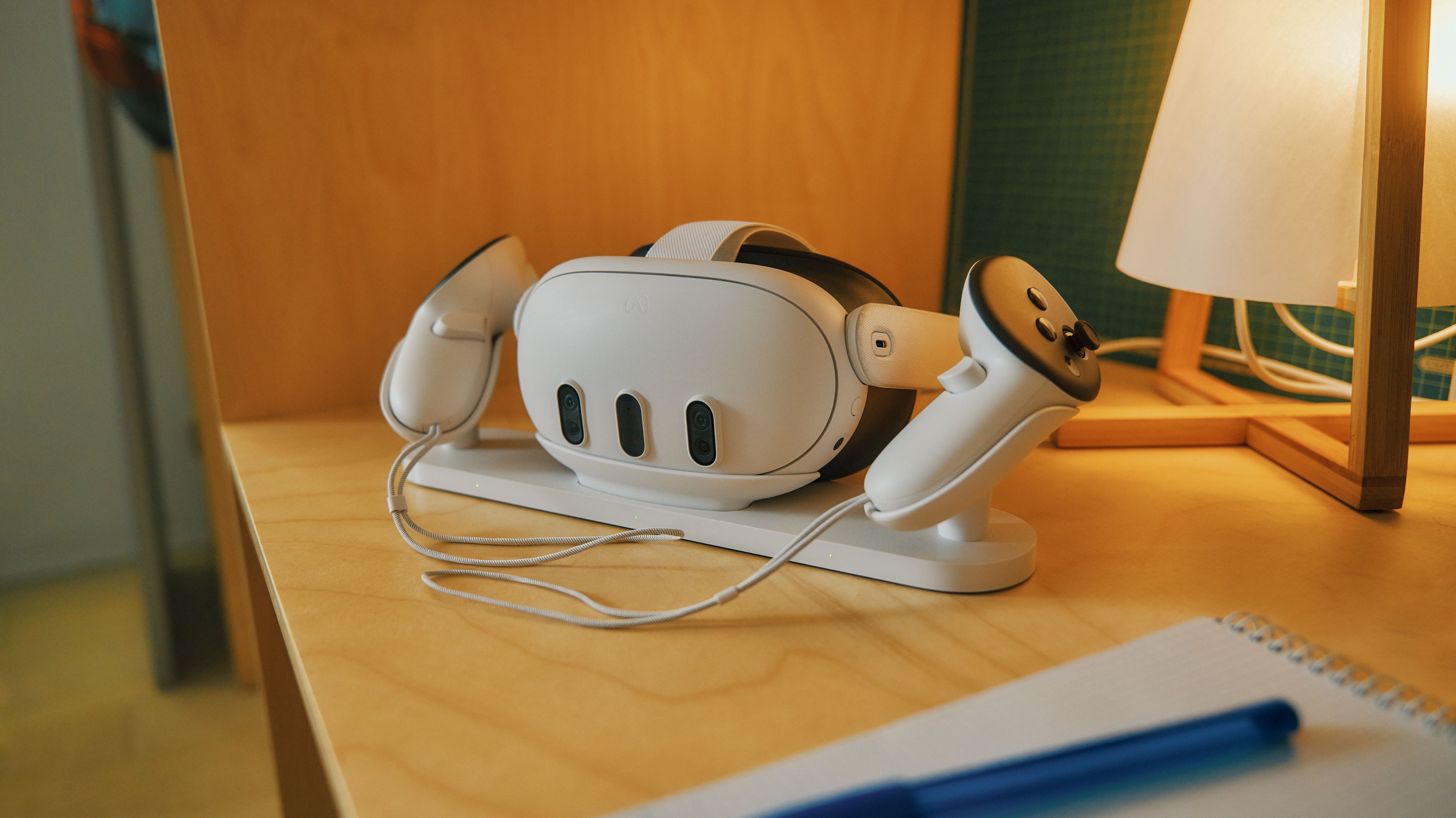 Meta Quest 3 and Controllers on their charging station that is on a wooden edge next to a lamp