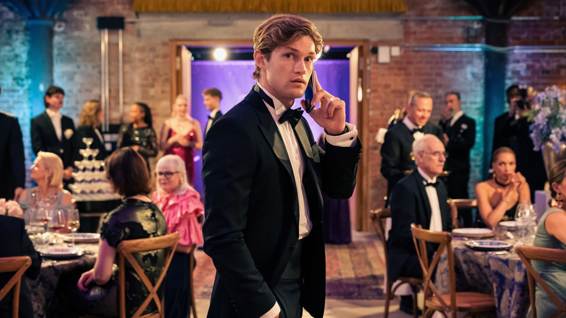 James holds a phone to his ear in the middle of a seated party