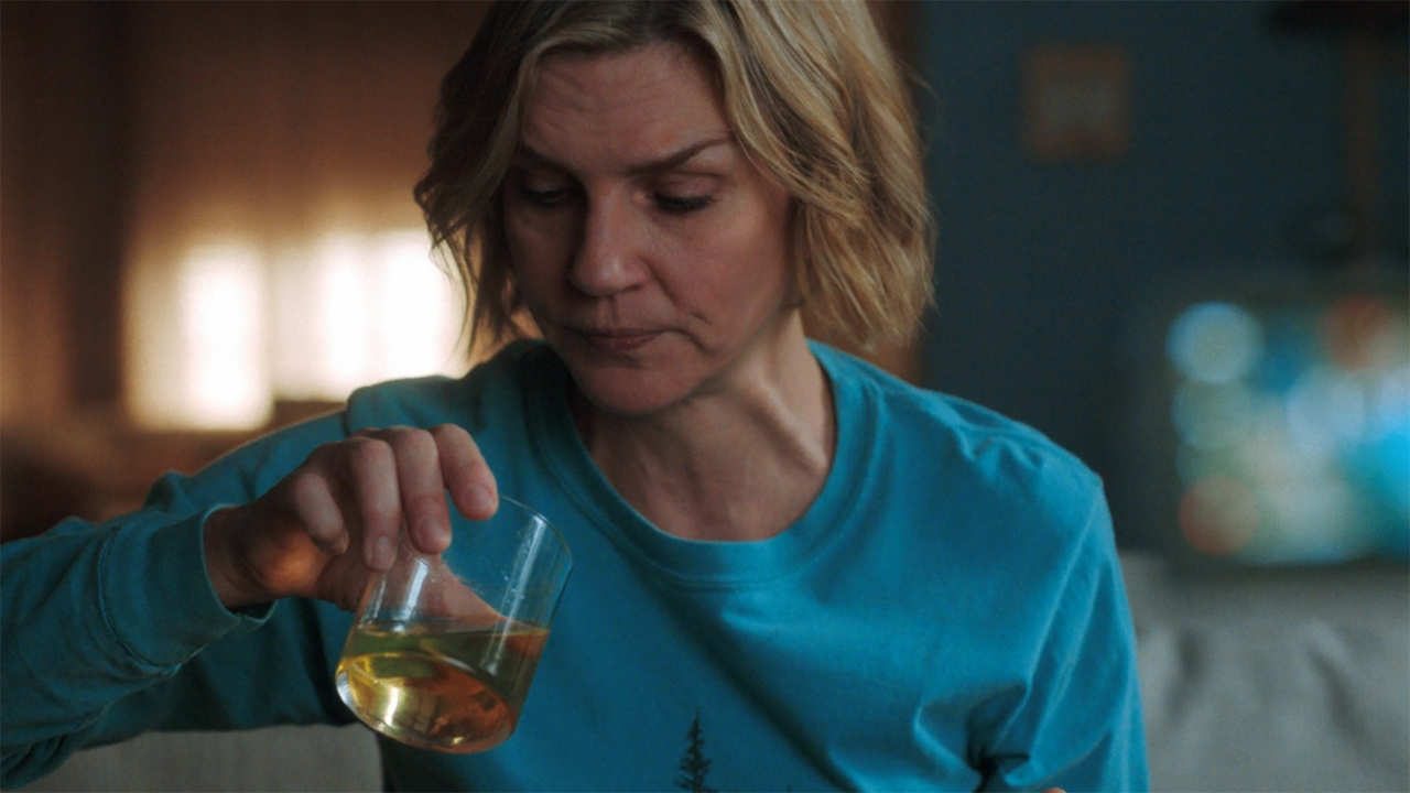 Carol sits down and looks at an orange liquid in a glass in Pluribus episode 5