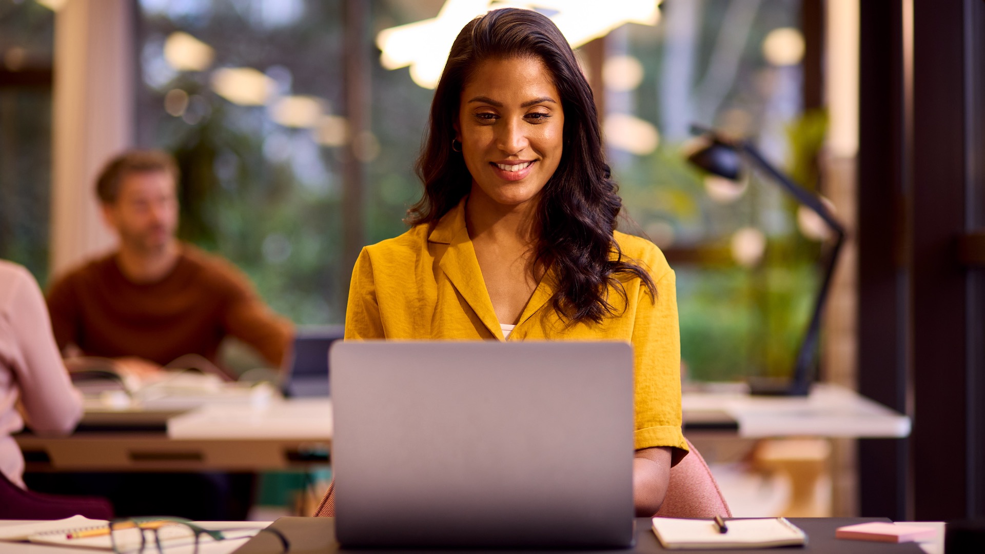 A young woman works on a Windows 11 laptop in a casual office space.