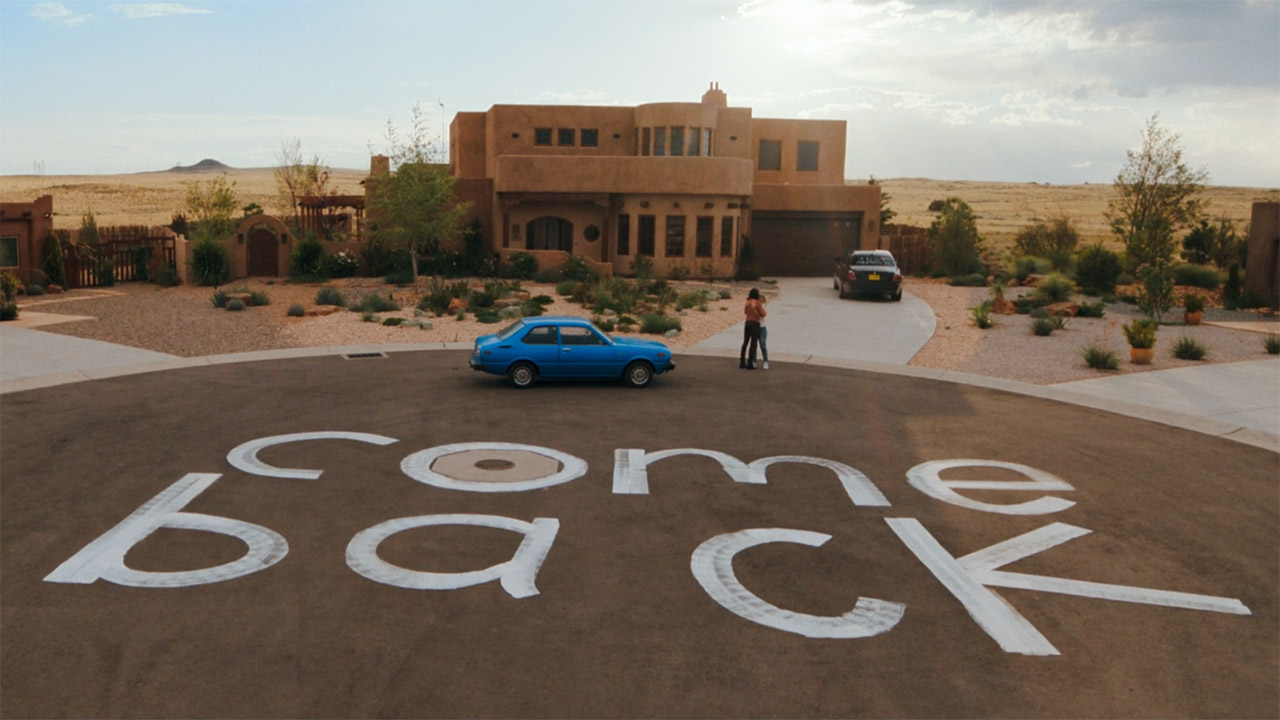 A wide shot of Zosia and Carol hugging near a giant painted 'come back' sign up Pluribus episode 7