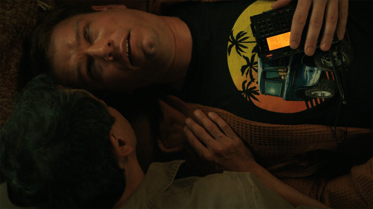 Manousos talking to a convulsed person with a portable radio lying on them in Pluribus episode 9