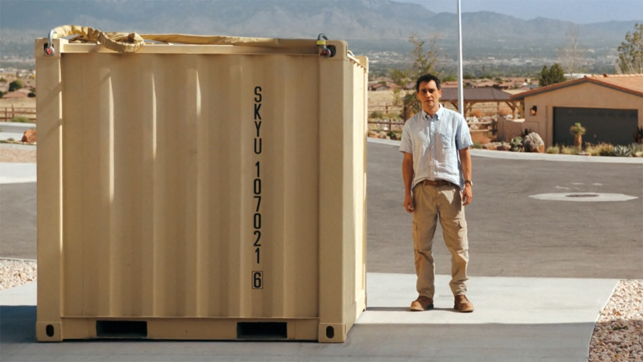 Manousos is standing next to a giant steel crate in Pluribus episode 9