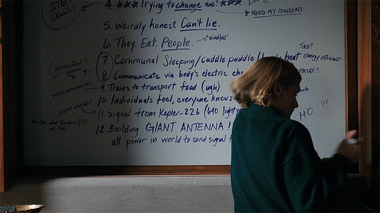Carol stands in front of a board with information about The Others written on it in Pluribus episode 8