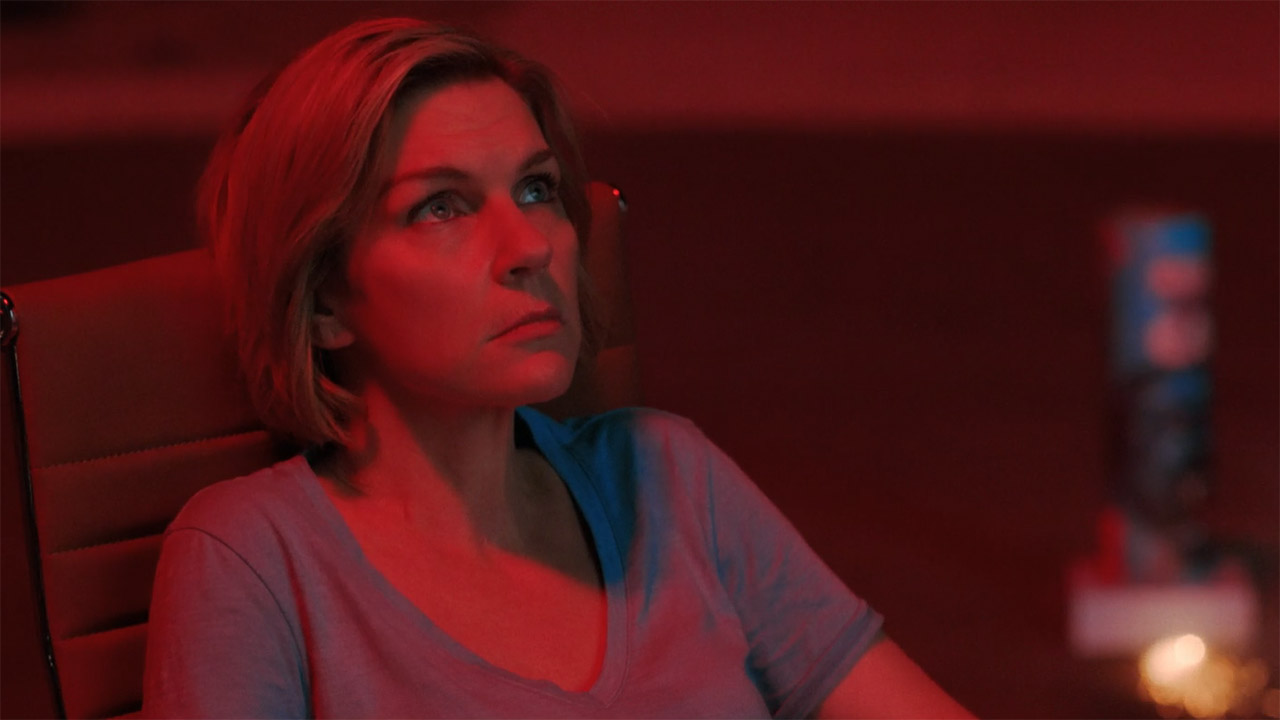 Carol sat down and bathed in red light from a fireworks display at night in Pluribus episode 7