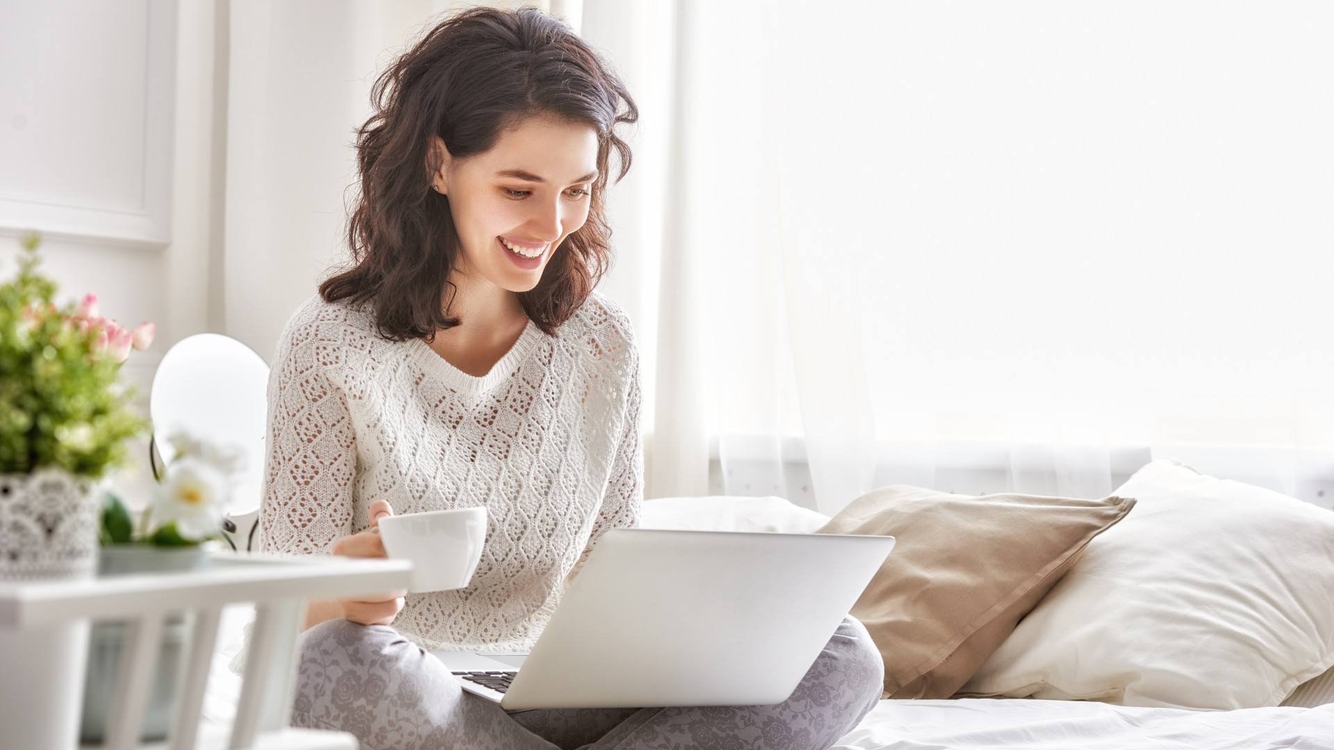 Happy woman sitting on a bed with a cup of coffee and a laptop
