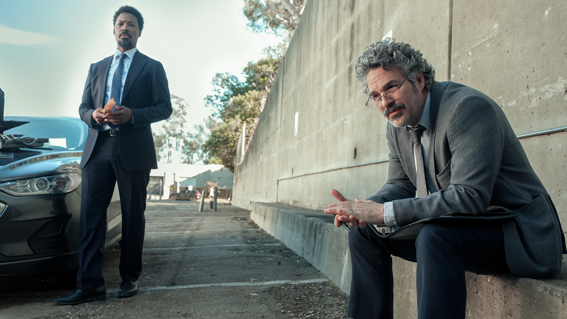 Detective Tillman stands next to a car while Detective Lubesnick sits on a nearby wall in Crime 101