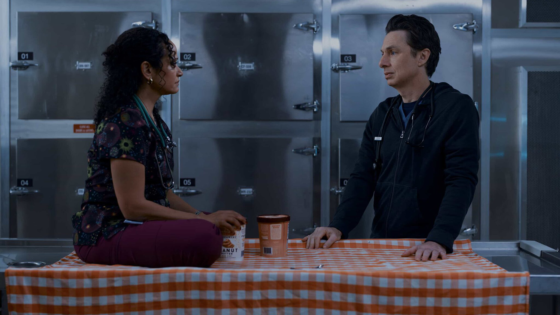 Carla sits astride a table that JD is standing next to in Scrubs Season 10 Episode 7