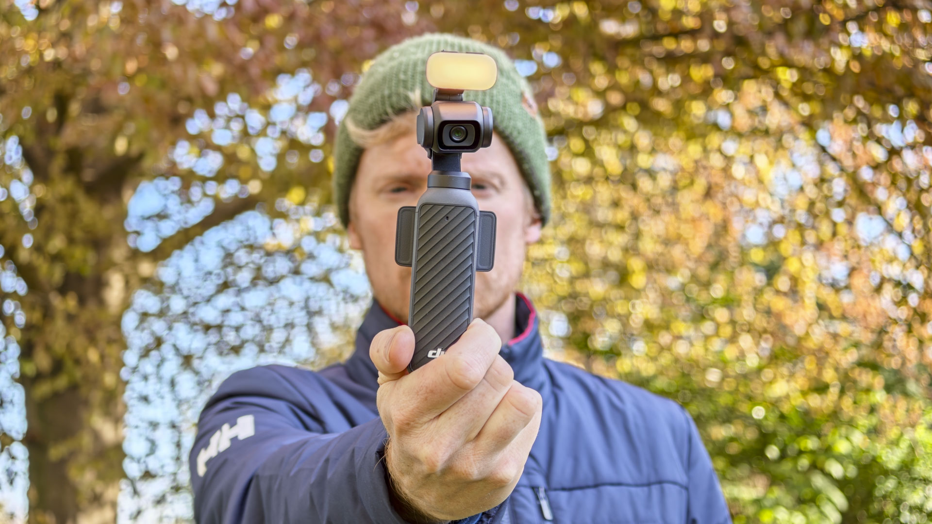 Man in beanie holding the DJI Osmo Pocket 4 vlogging camera with its magnetic fill light, an autumn tree behind