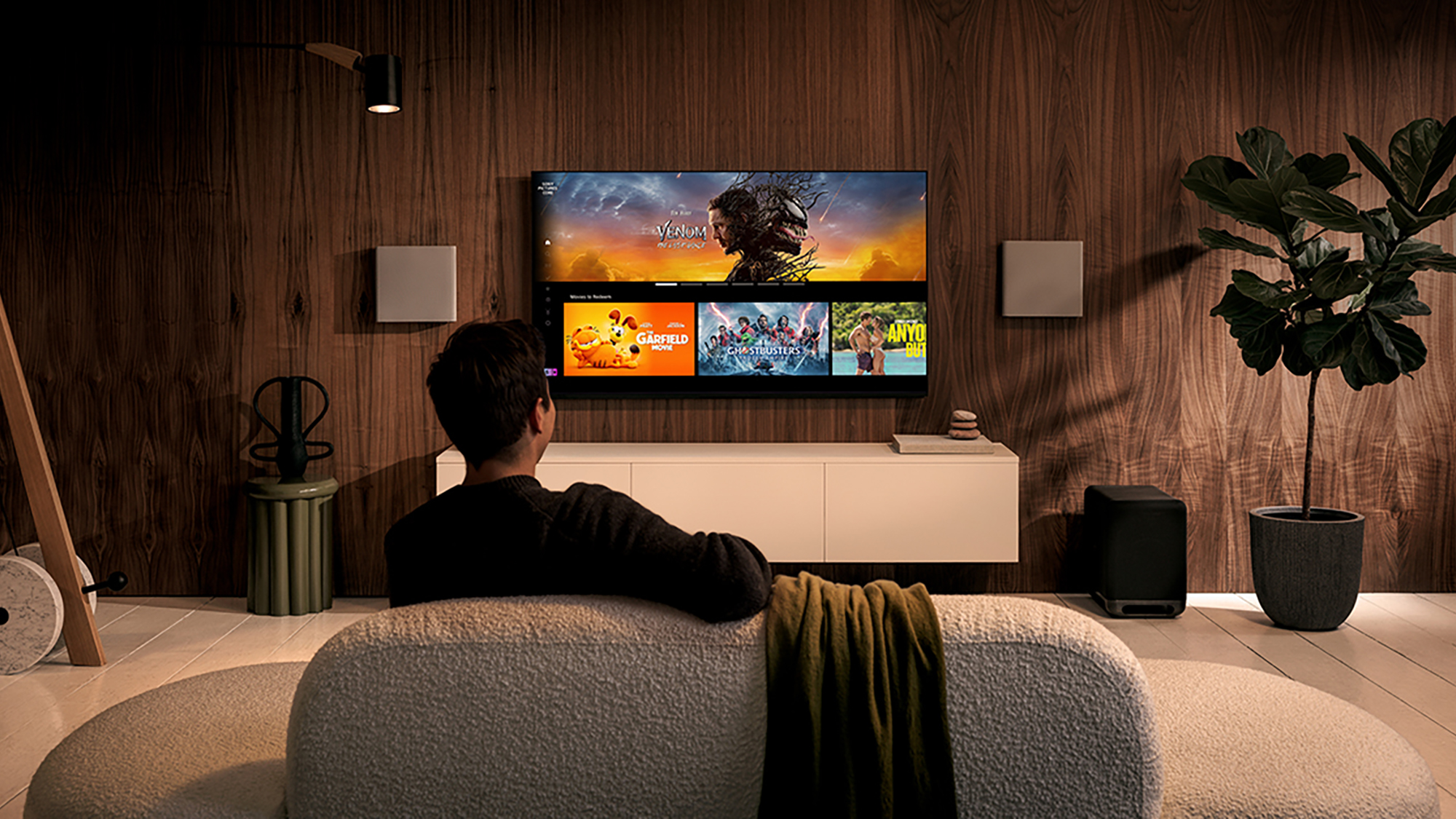 Rear view of a man sitting on a couch looking at a TV showing thumbnails of selected content