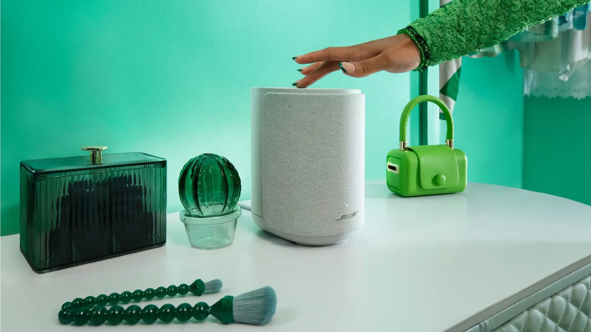 Bose Lifestyle Ultra Speaker on a white surface in front of a green wall. There are green ornaments on the white surface, and a person's hand is touching the top of the speaker.