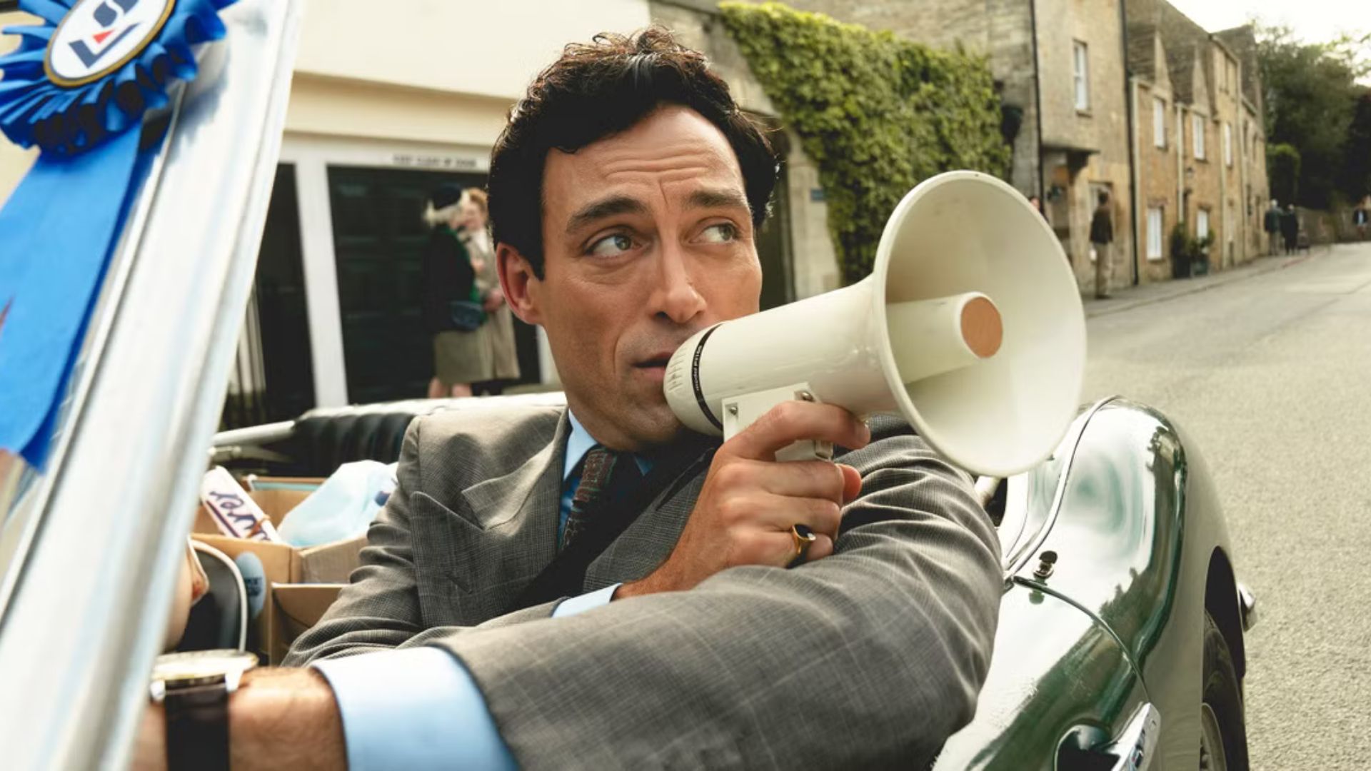 Rupert Campbell-Black speaks through a megaphone while driving a convertible