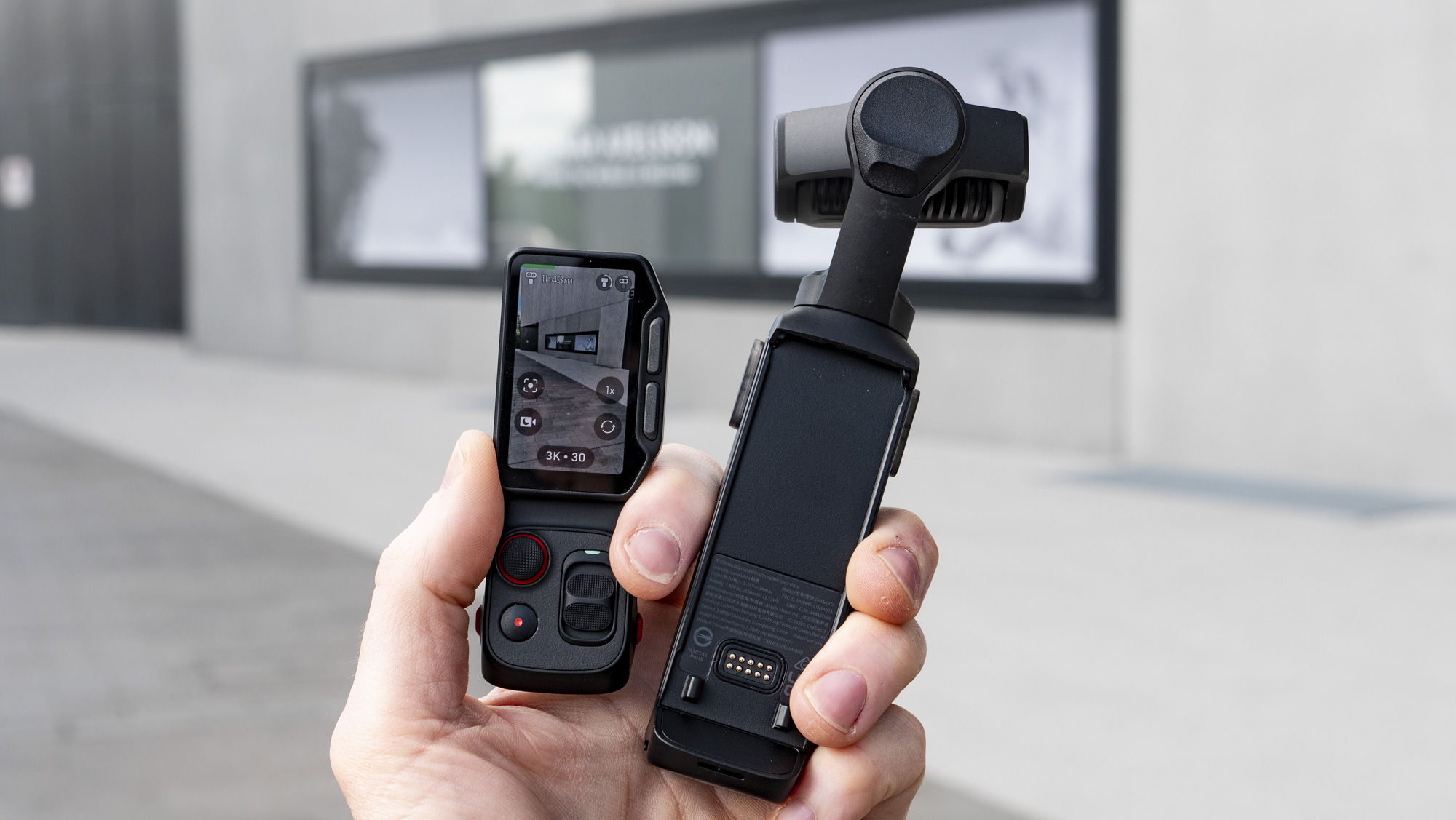 Hand holding the Insta360 Luna Ultra vlogging camera and its screen, outdoors in a gray urban scene