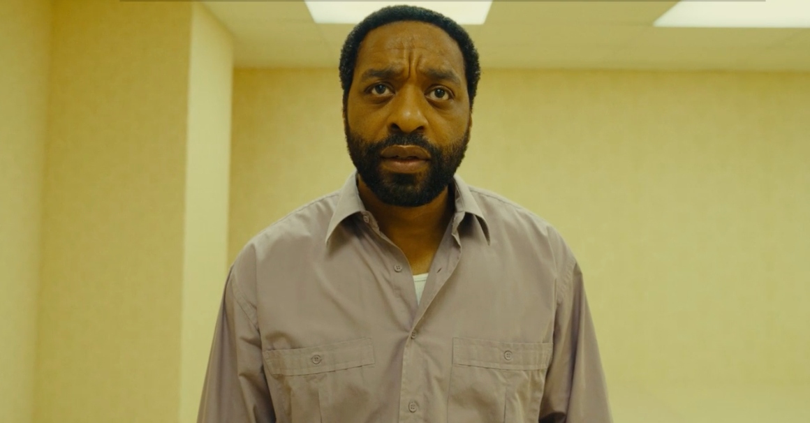 Chiwetel Ejiofor stands in the back rooms, where he is surrounded by pale yellow walls and fluorescent lights.