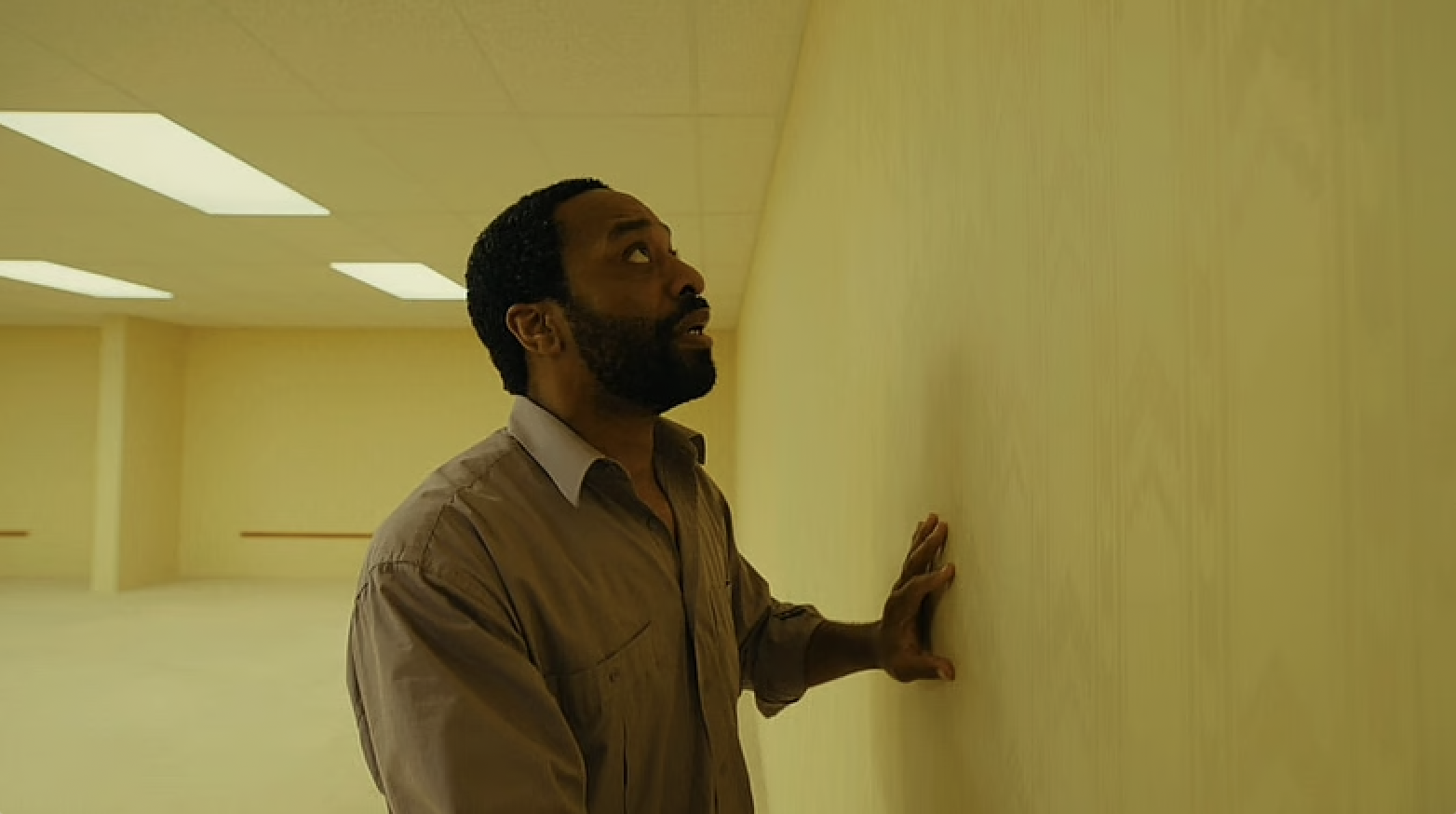 Chiwetel Ejiofor in Backrooms where he is in an empty yellow room. He touches a wall and looks scared.