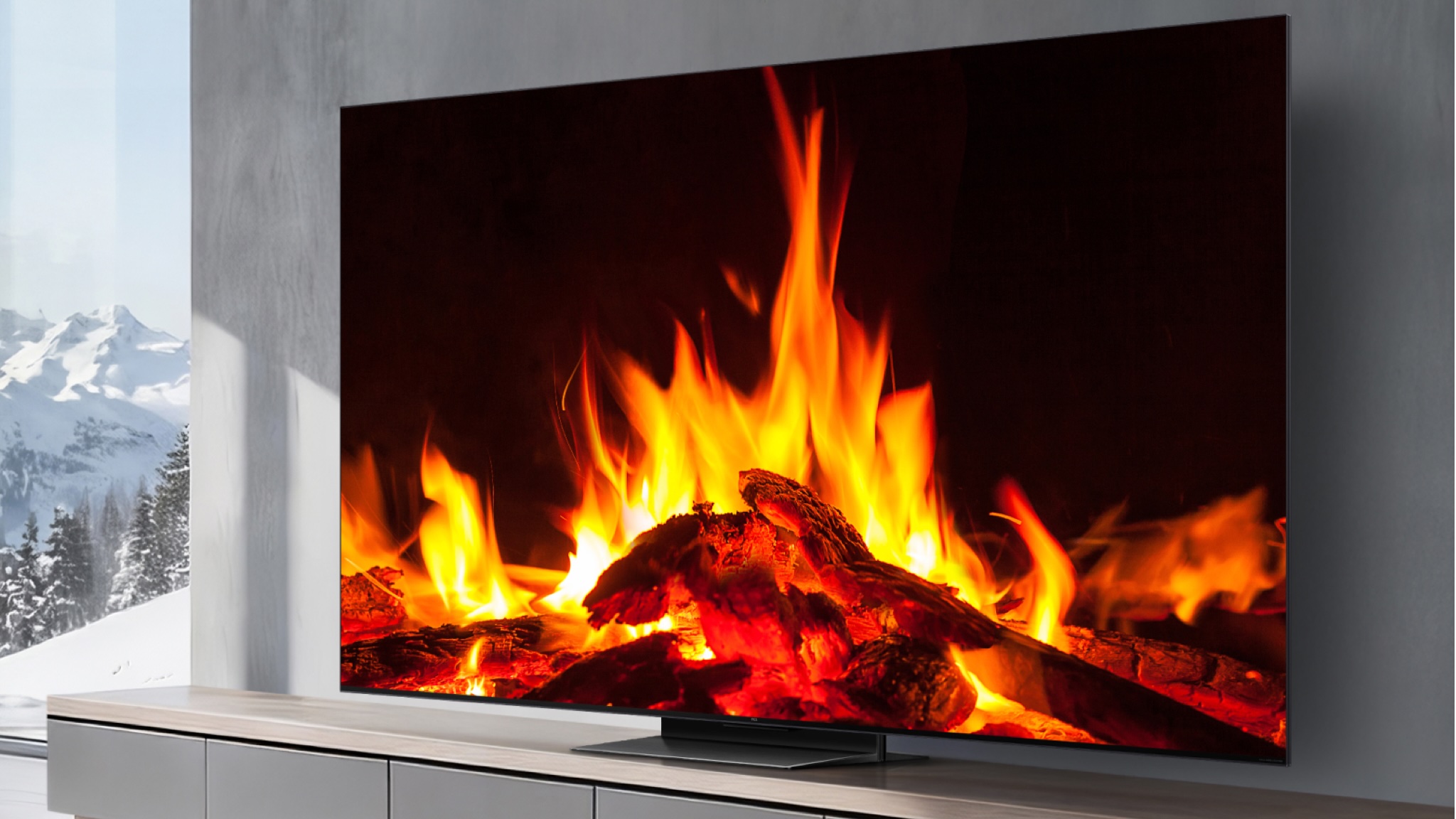 A TCL C7L TV showing a wood stove. It is in a gray decorated room with snowy mountains visible through a full height window to the left of the TV unit