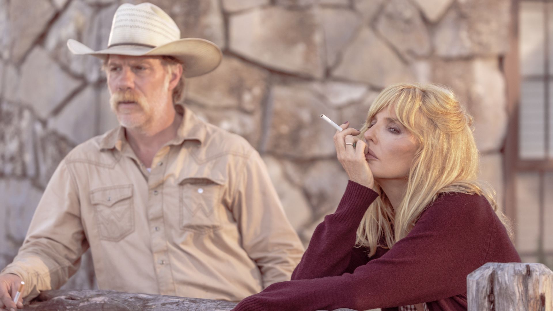 Beth smokes as she leans against a wooden plank with Zachariah standing behind