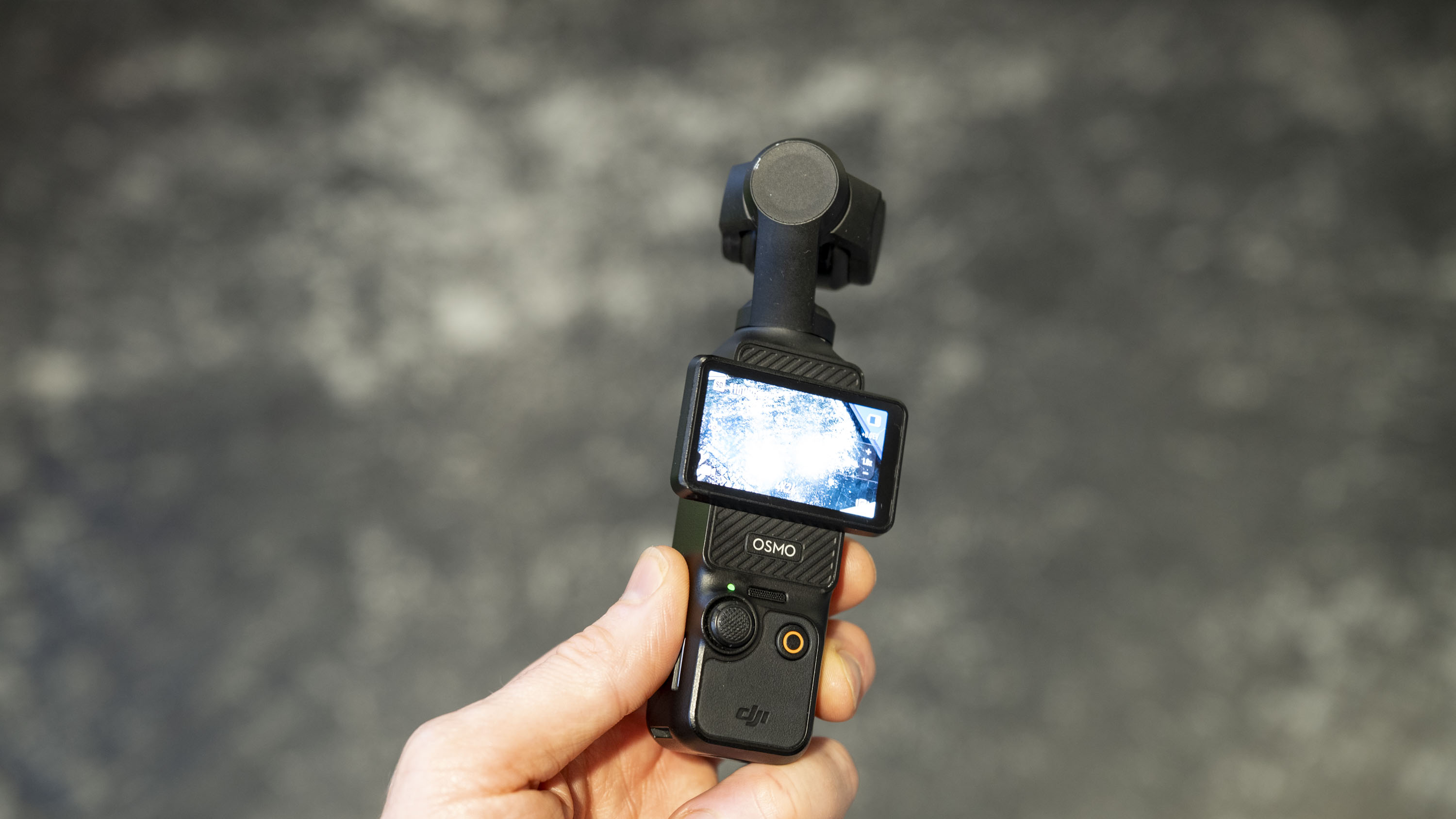 DJI Osmo Pocket 3 vlogging camera in the user's hand, gray floor in the background