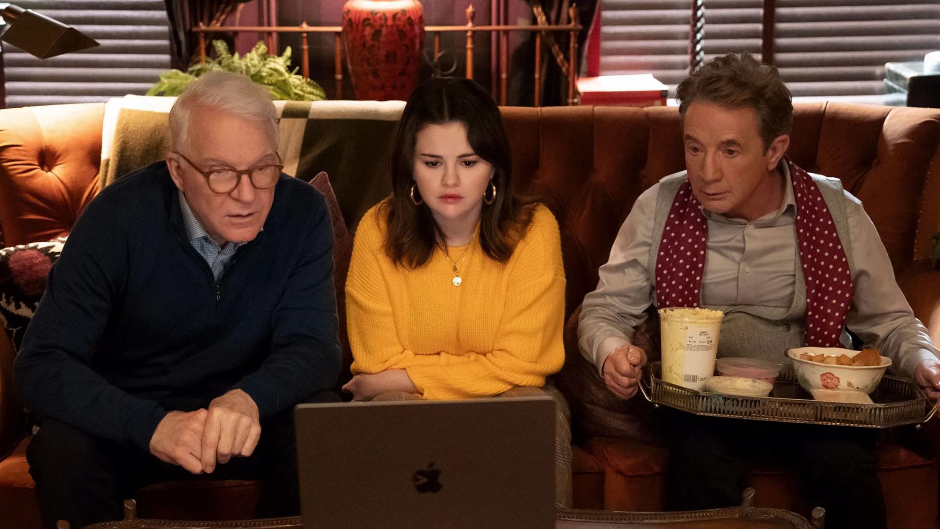 Steve Martin, Selena Gomez and Martin Short sit on a sofa looking at a laptop with a tray of food on Short's lap