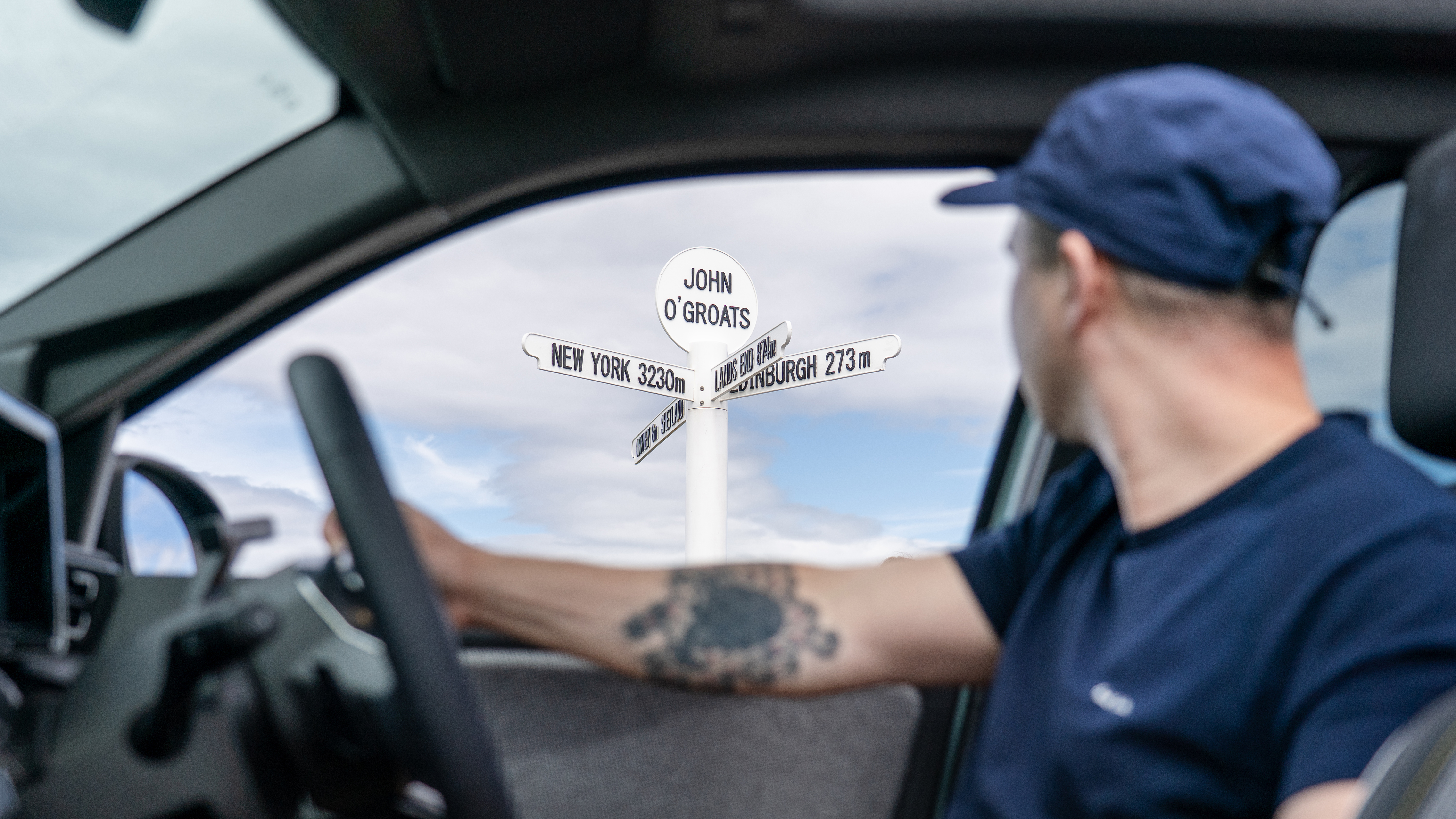 A man looks out of the window of an electric car at a road sign for John o'Groats