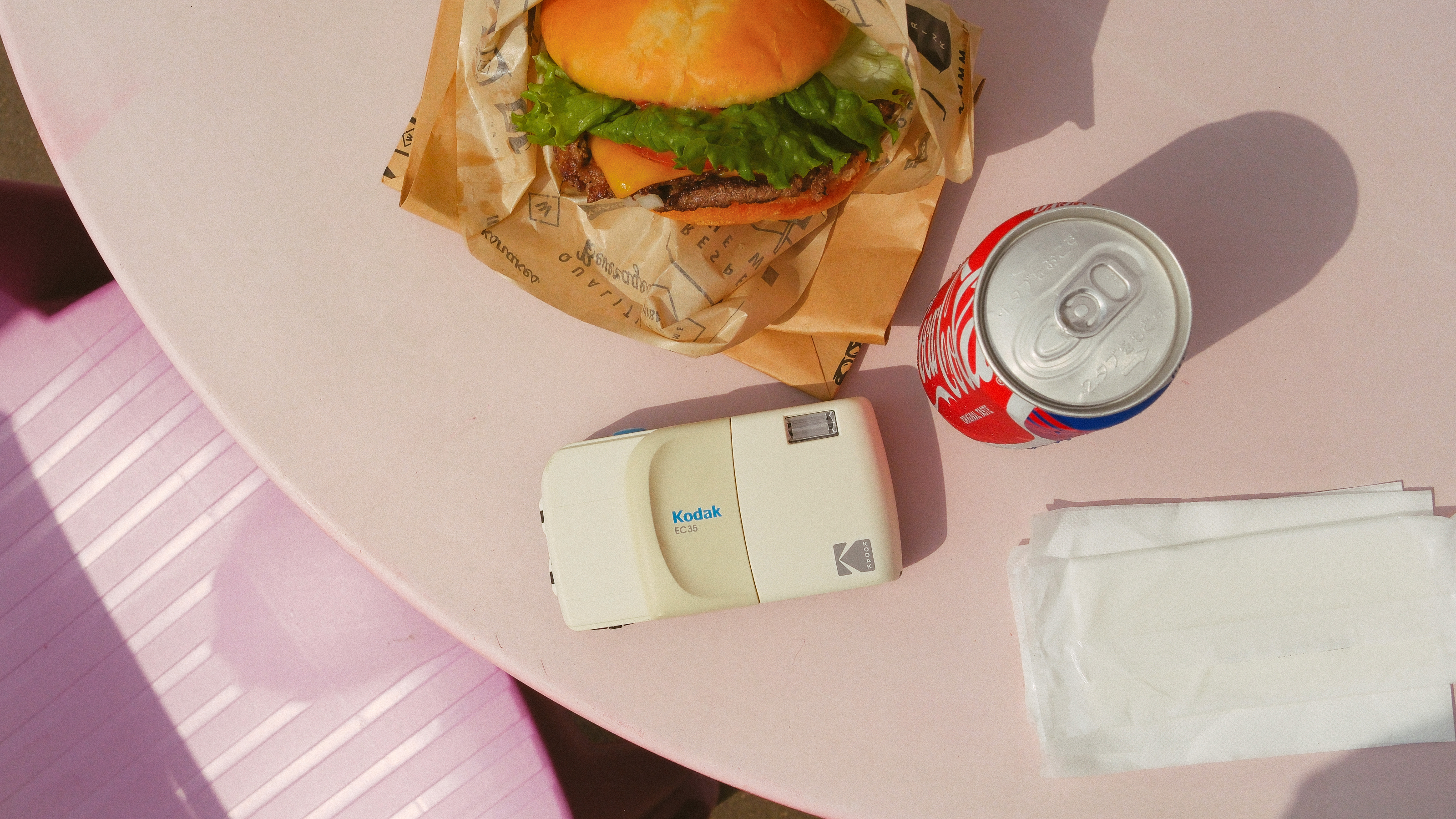 Photograph of the Kodak EC35 film camera in white on a table next to a burger and a can of coke