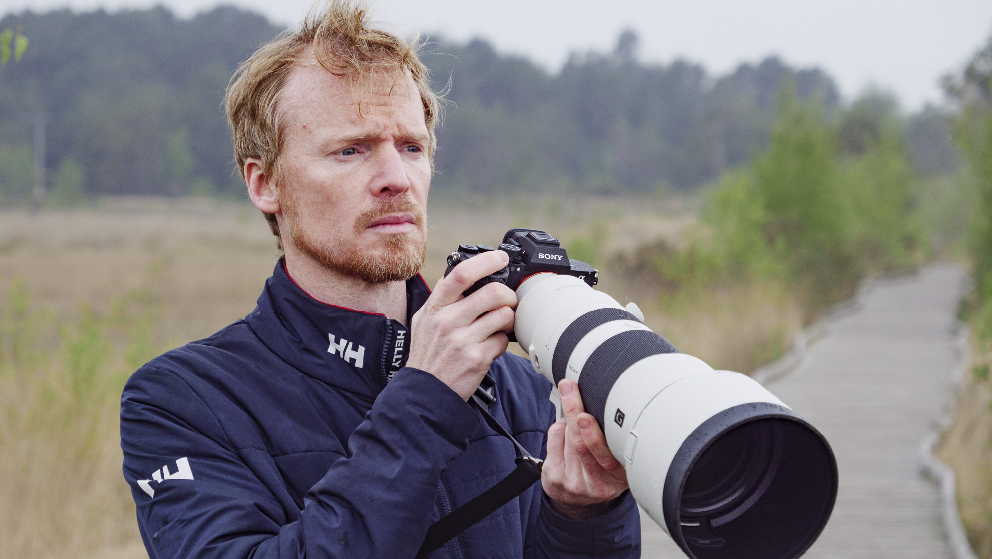 Man holding Sony A7R VI camera with FE 400-800mm F6.3-8 G OSS super telephoto zoom lens attached, standing on boardwalk in nature reserve looking for birds