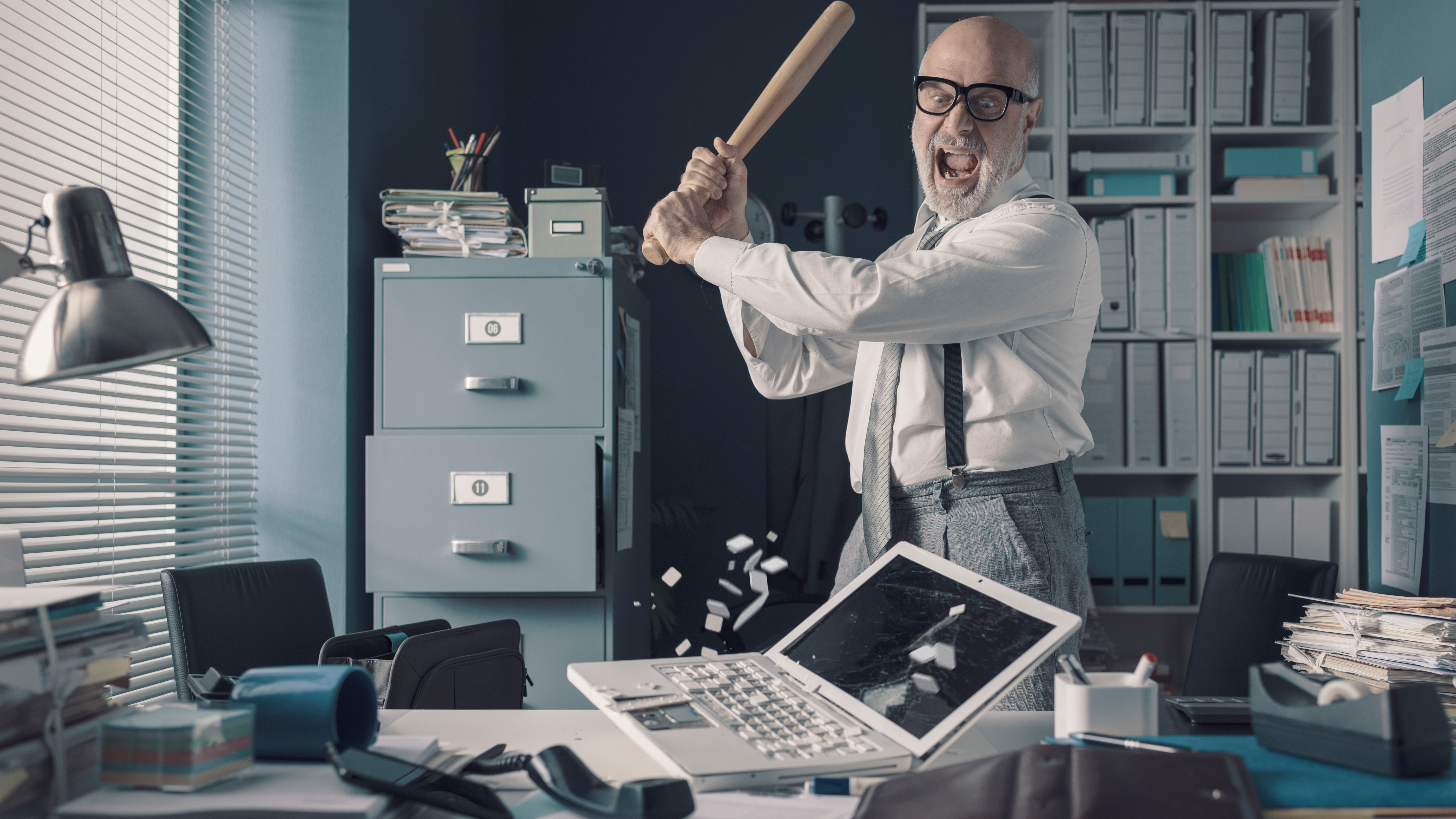 Angry businessman destroys his desk and laptop with a baseball bat